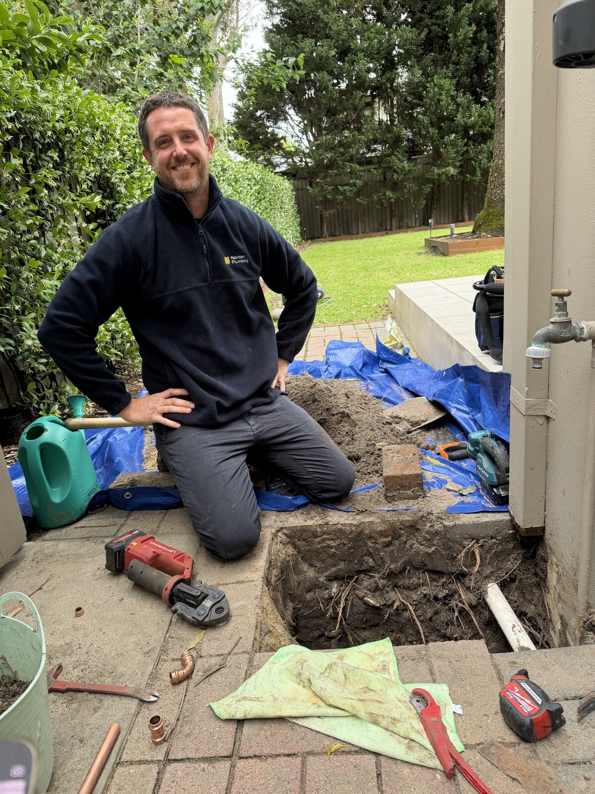 Adam Norton smiling beside the finished burst water pipe repair in a Maroubra garden, with the press tool and new copper fittings on the pavers