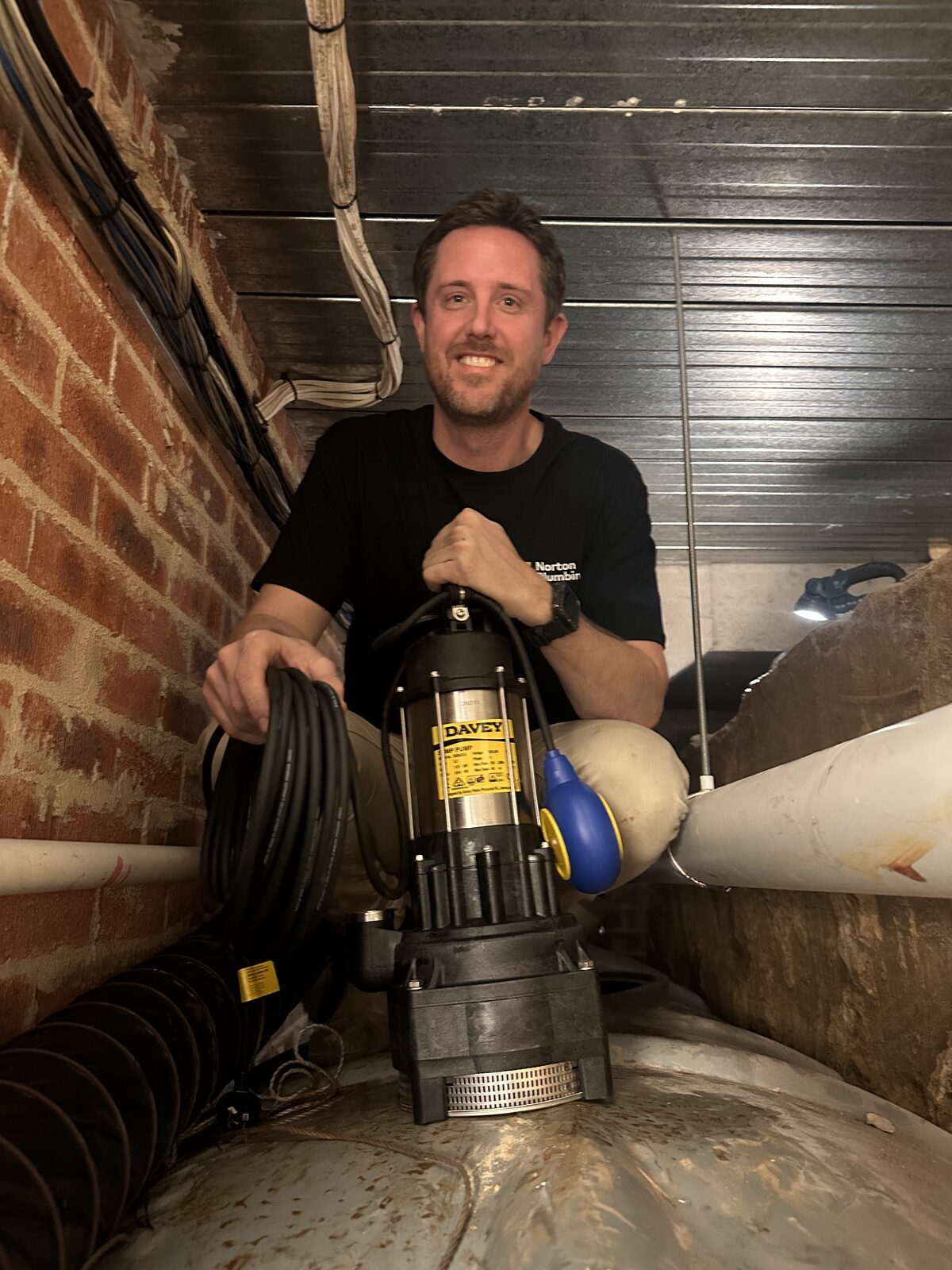 Adam Norton from Norton Plumbing holding a new Davey submersible pump with a float switch in the subfloor of a Dover Heights house during a rainwater pump replacement