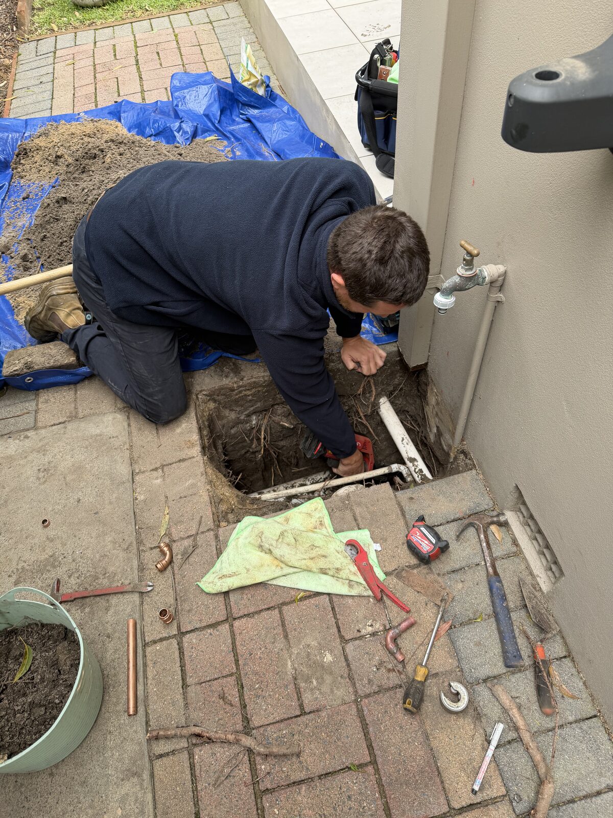 Adam Norton kneeling on a tarp beside a house in Maroubra, digging out wet soil under lifted brick pavers to expose a burst copper water pipe