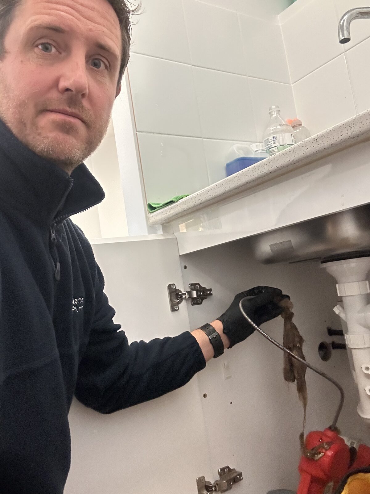 Adam Norton from Norton Plumbing working at an open under sink cabinet at a Maroubra salon with a drain machine set up to clear the blocked drain