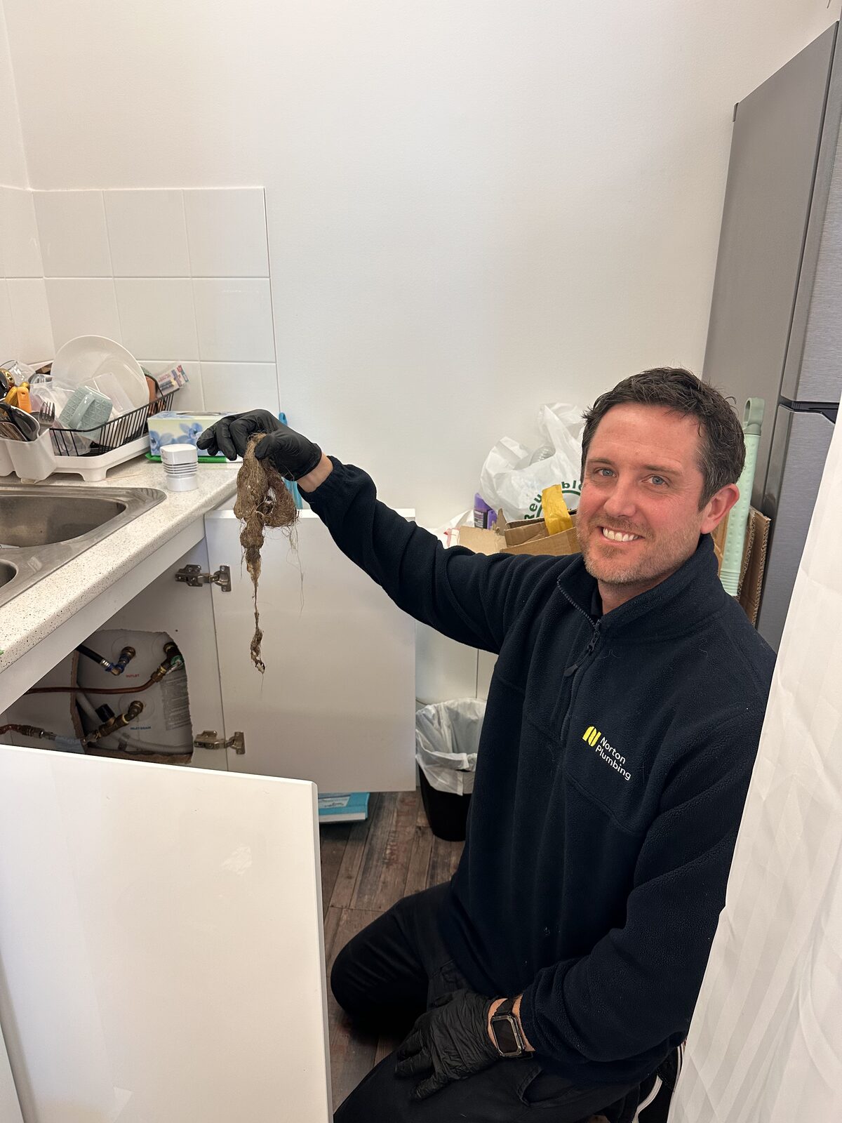 Adam Norton from Norton Plumbing kneeling beside an open under sink cabinet at a Maroubra hairdressing salon, holding a large mass of matted hair cleared from the blocked drain
