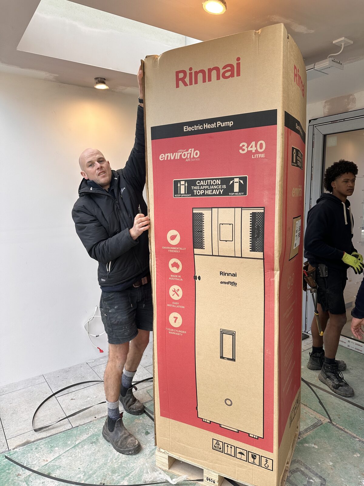 Adam Norton and Greg from Norton Plumbing moving a boxed Rinnai Enviroflo 340 litre heat pump hot water system into a Maroubra home