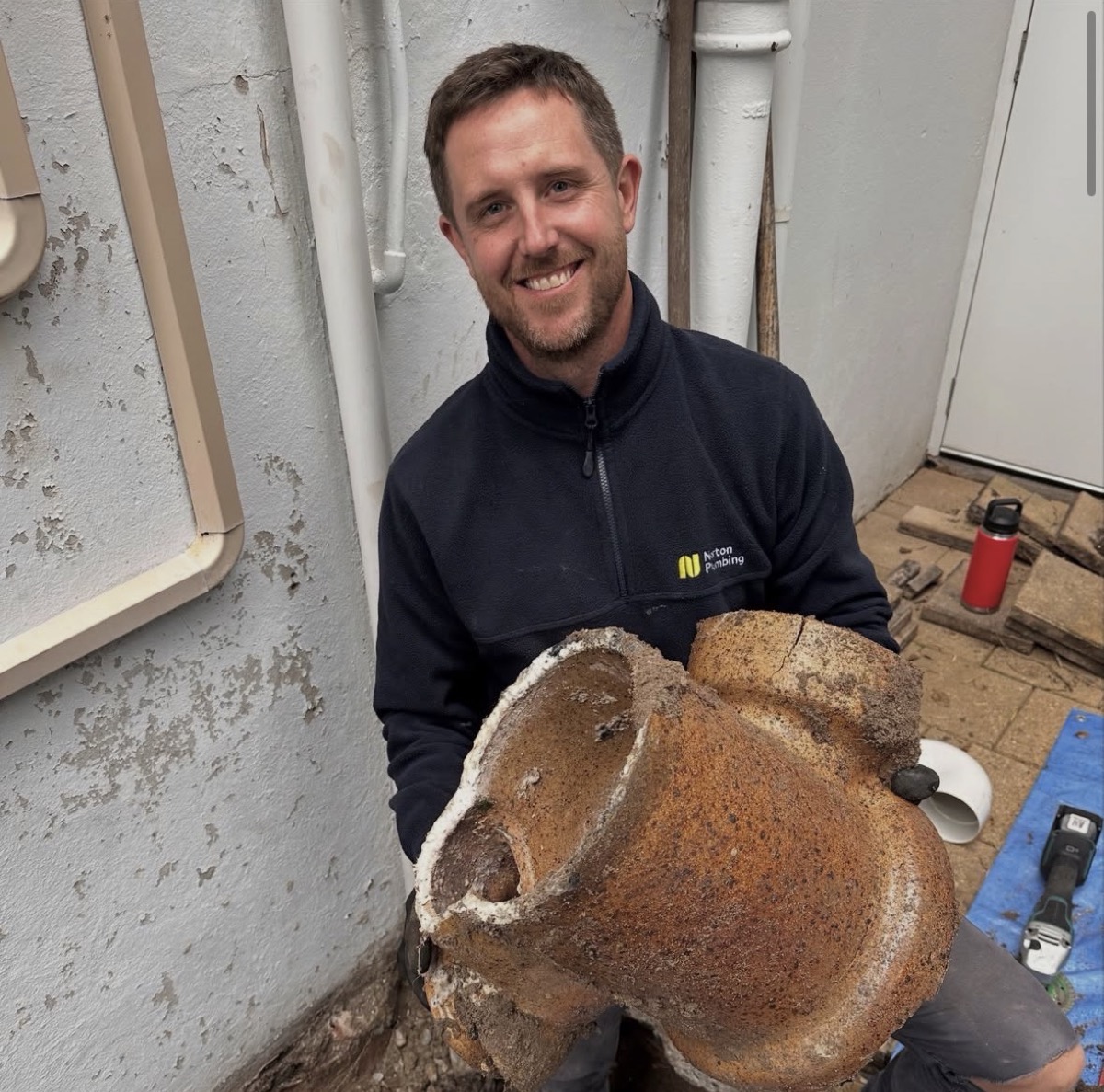 Adam Norton from Norton Plumbing holding the cracked terracotta drain junction removed from the 1930s blocked drain at Mount Street Coogee, smiling at camera