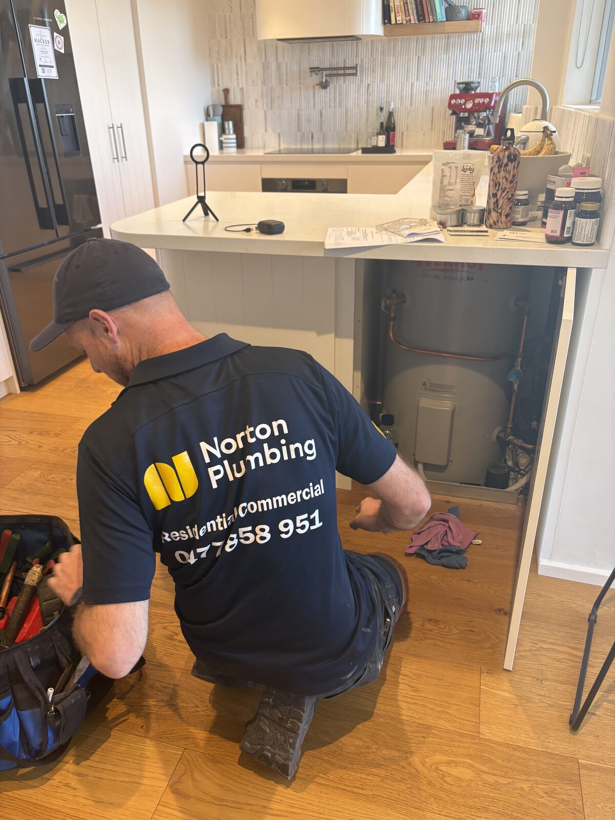 Adam Norton of Norton Plumbing kneeling at an open kitchen island cupboard containing an electric storage hot water tank at a Coogee home