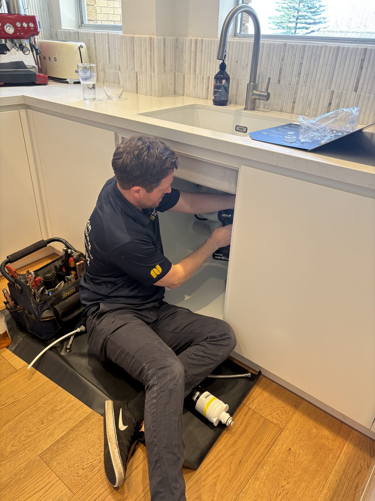 Adam Norton from Norton Plumbing sitting on a drop sheet fitting a Puretec Z7 under-sink water filter inside the sink cupboard of a Coogee apartment kitchen