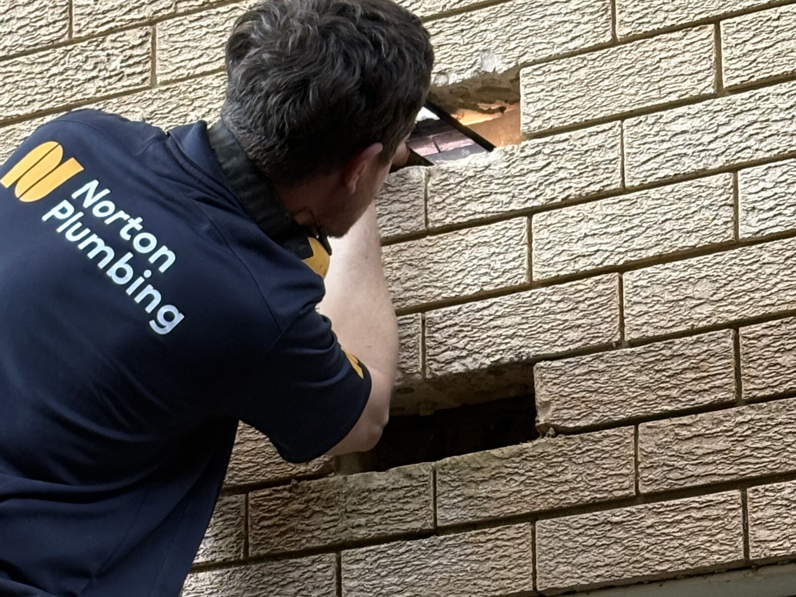 Adam Norton from Norton Plumbing holding a phone into an opening cut in the brick wall of a Coogee block of flats to see the burst pipe inside the cavity