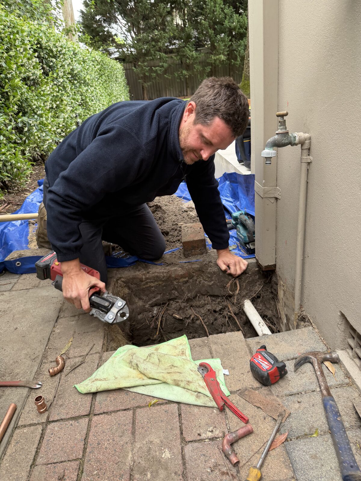 Adam Norton kneeling over an excavated hole beside a house in Maroubra, using a press tool to join new copper pipe to the repaired water line under the pavers