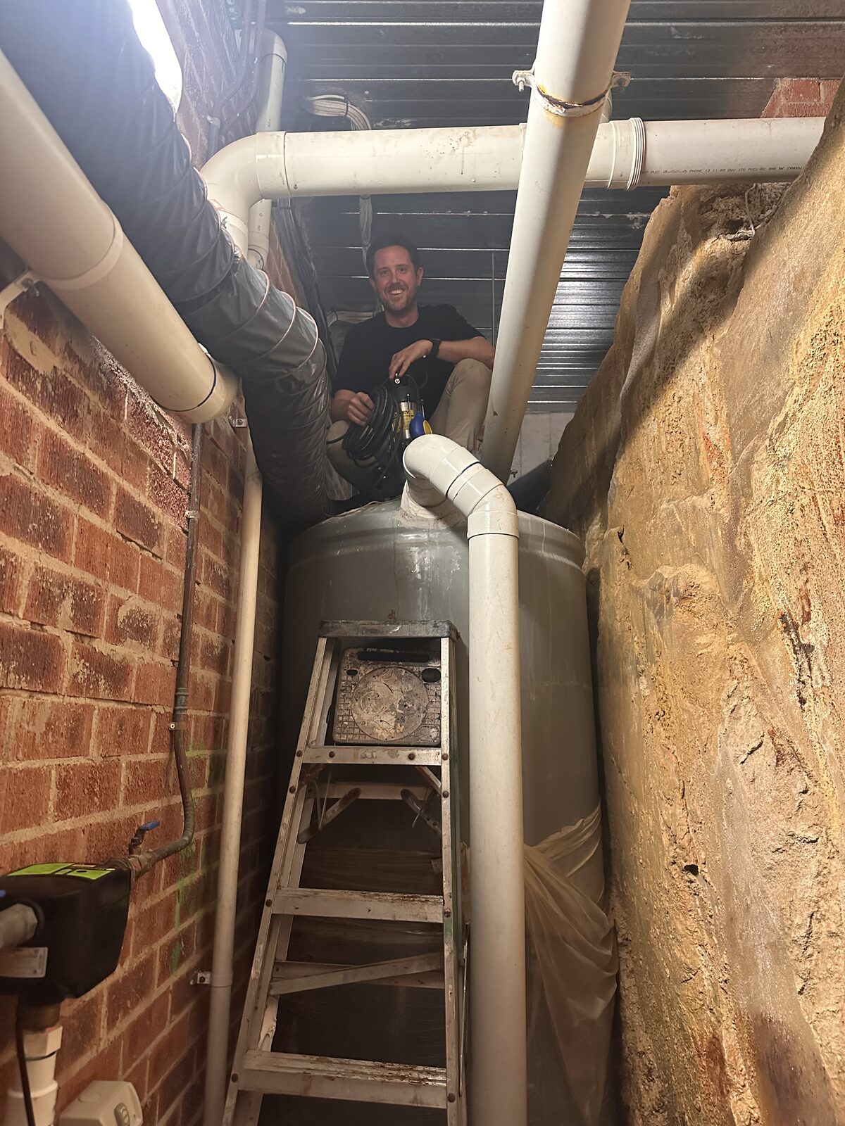 Rainwater tank in the narrow subfloor void of a Dover Heights house, wedged between a brick wall and a cut sandstone rock face, with a ladder providing access over the top of the tank