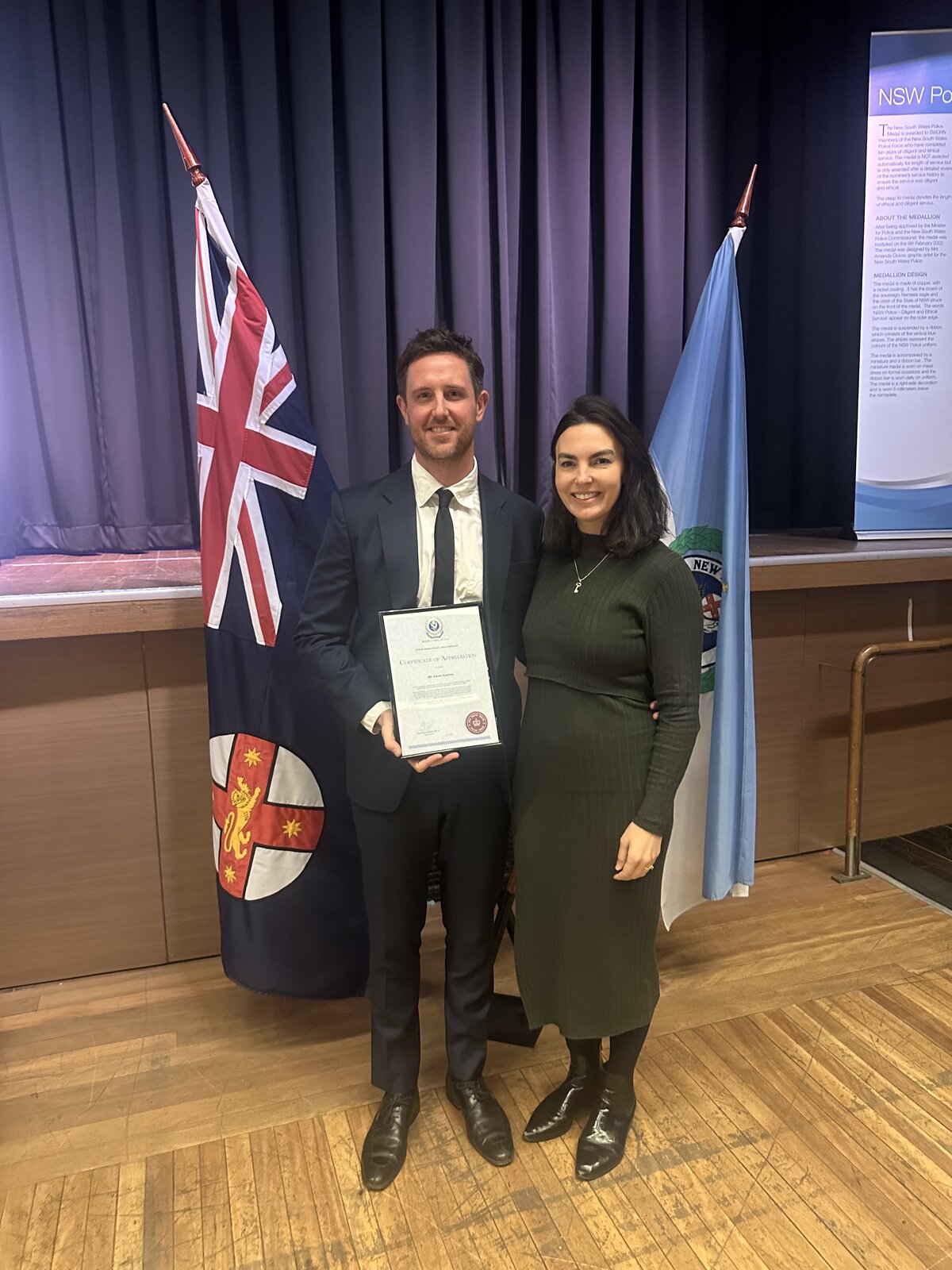 Adam and Rebecca Norton holding Adam's NSW Police Certificate of Appreciation, standing in front of the NSW and Australian flags