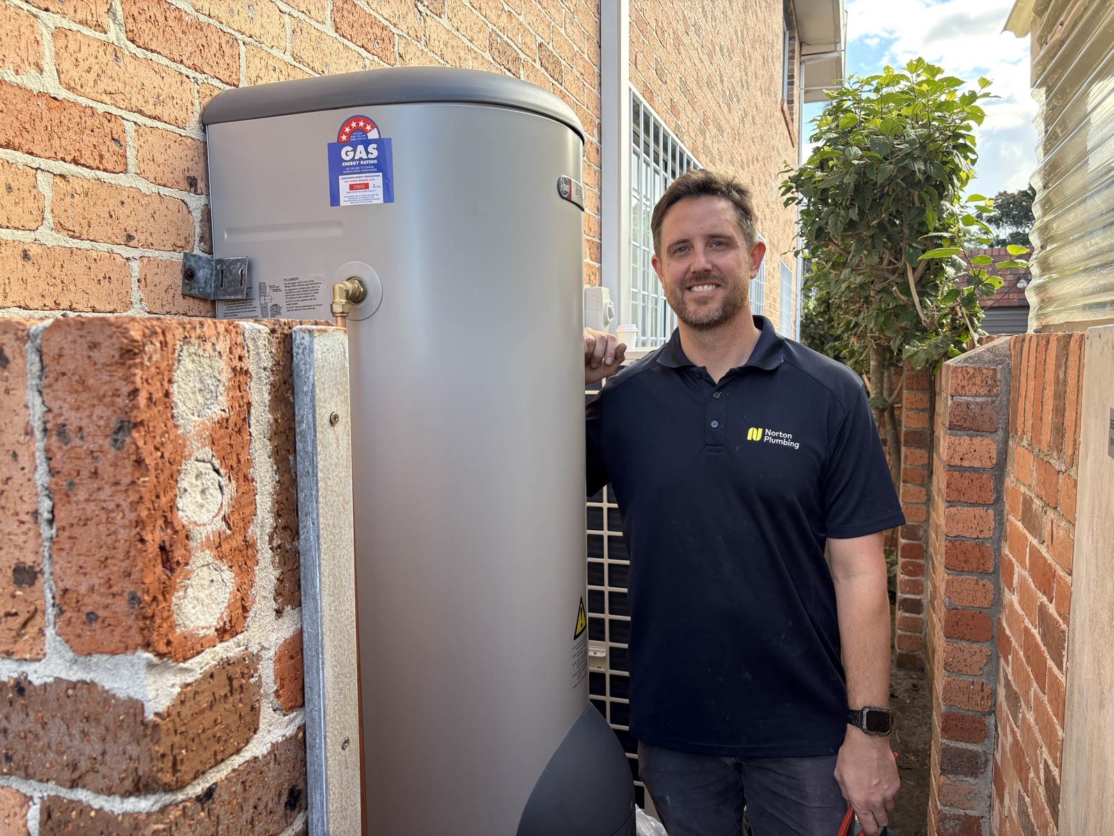 Adam Norton from Norton Plumbing standing beside a newly installed Rheem gas hot water system in a brick side passage in Maroubra