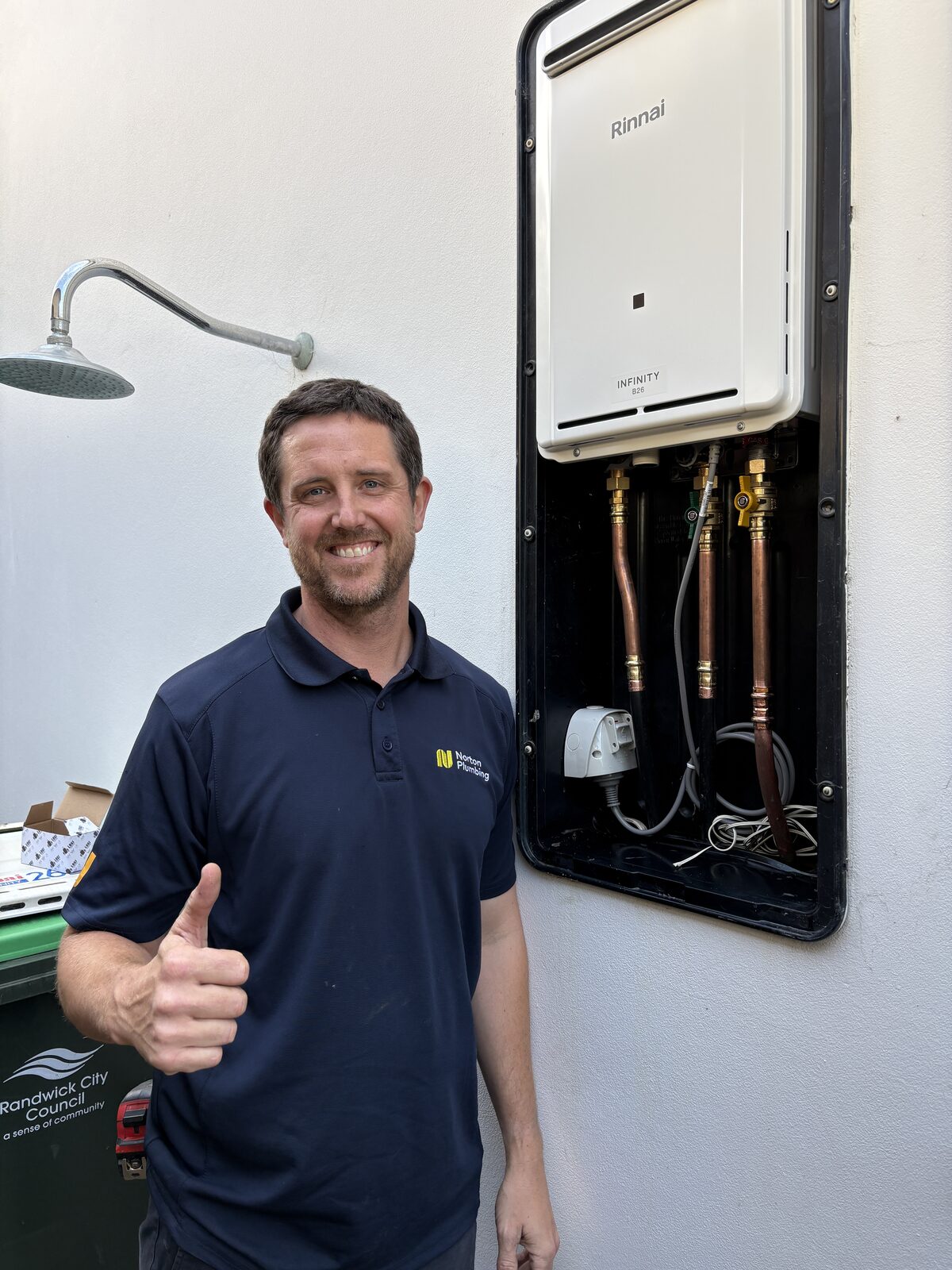 Adam from Norton Plumbing giving a thumbs up beside a new Rinnai Infinity B26 gas instantaneous hot water system in a wall recess box at a Clovelly house