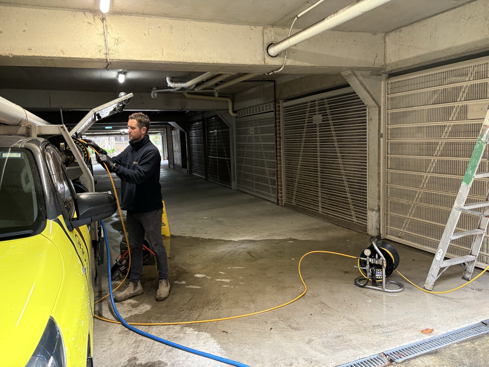 Adam Norton setting up high-pressure jetting equipment in the underground car park of a Rose Bay apartment building, with CCTV inspection reel on the ground beside the yellow Norton Plumbing truck