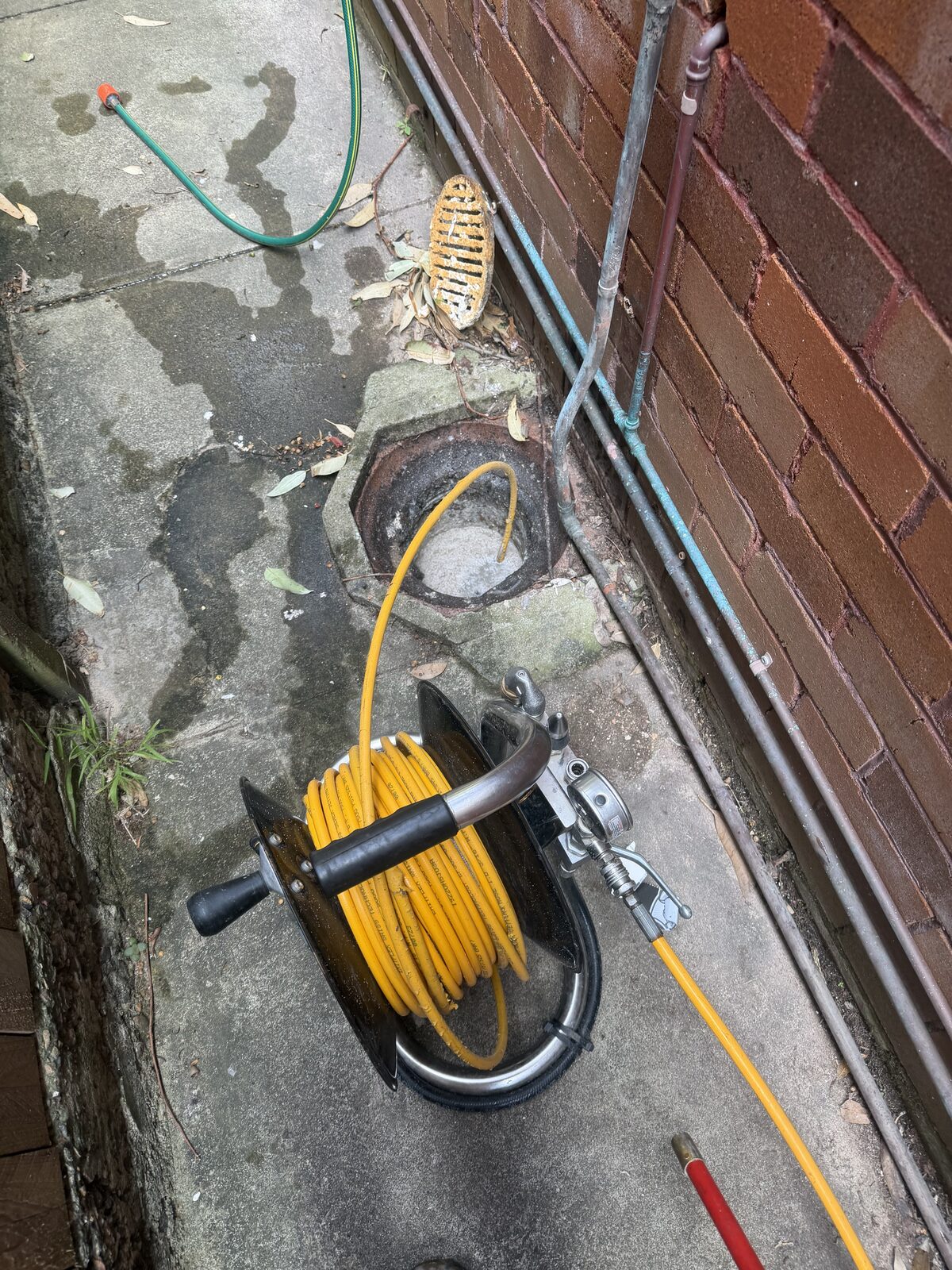 Close up of a drain jetter reel with yellow high pressure hose fed into an open sewer gully in a concrete side passage at a Bondi strata block