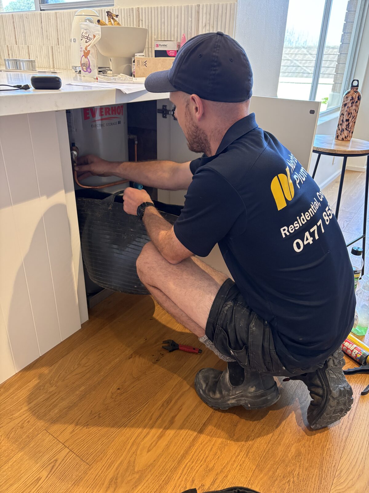 Adam Norton working on the pipework of an Everhot electric storage tank inside a kitchen island cupboard at a Coogee home, with a drain tub in place