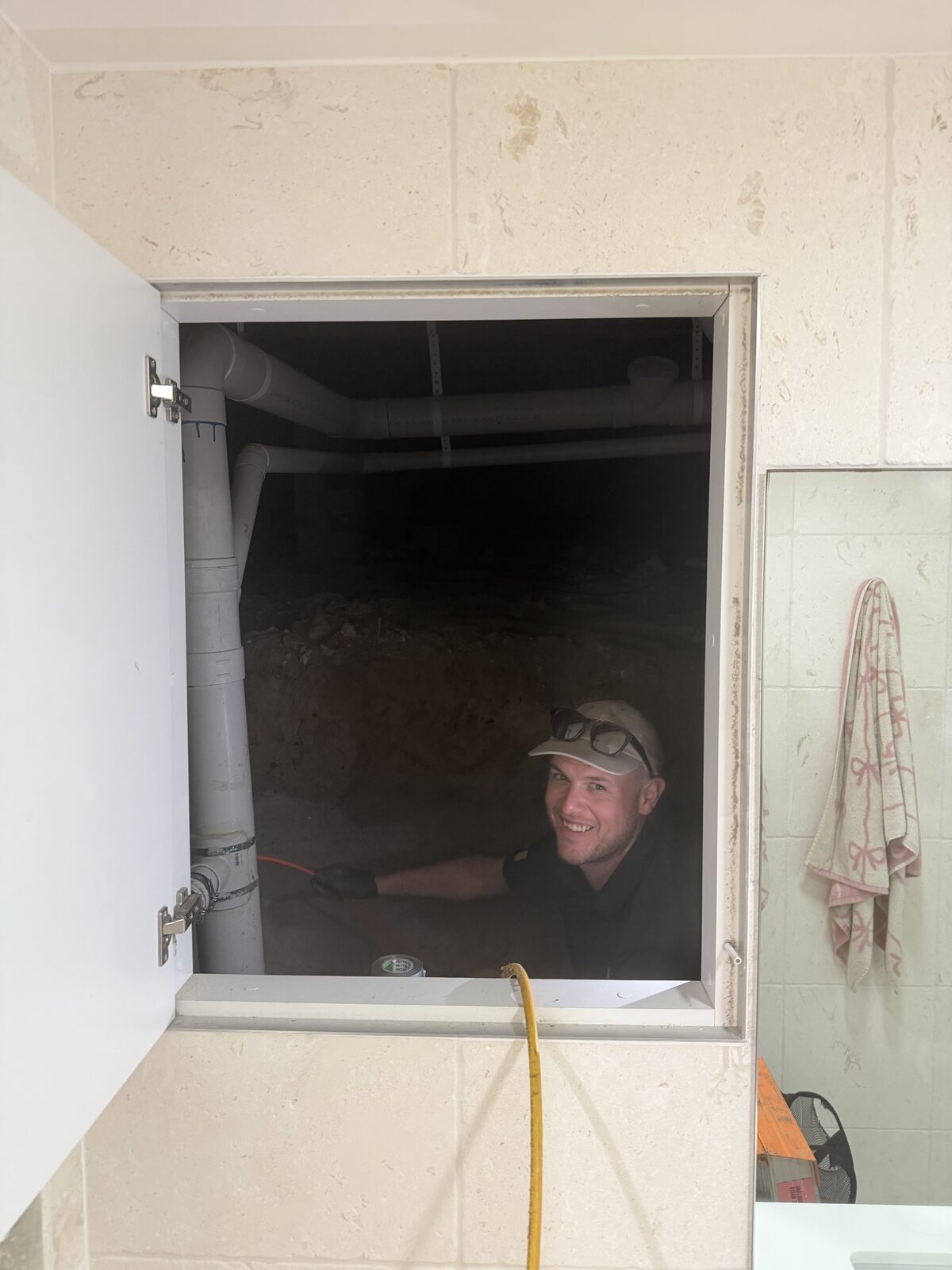 Greg from Norton Plumbing in the subfloor of a Bronte home, seen through the access panel in the bathroom wall, clearing a blocked basin drain with the jetter hose