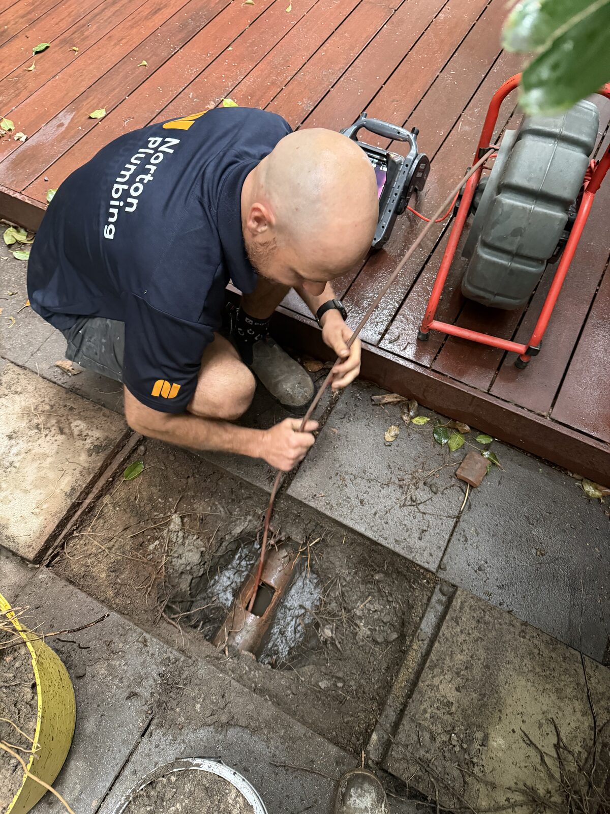 Greg from Norton Plumbing feeding a CCTV drain camera into an opened terracotta stormwater pipe under lifted pavers next to a timber deck at a Double Bay home
