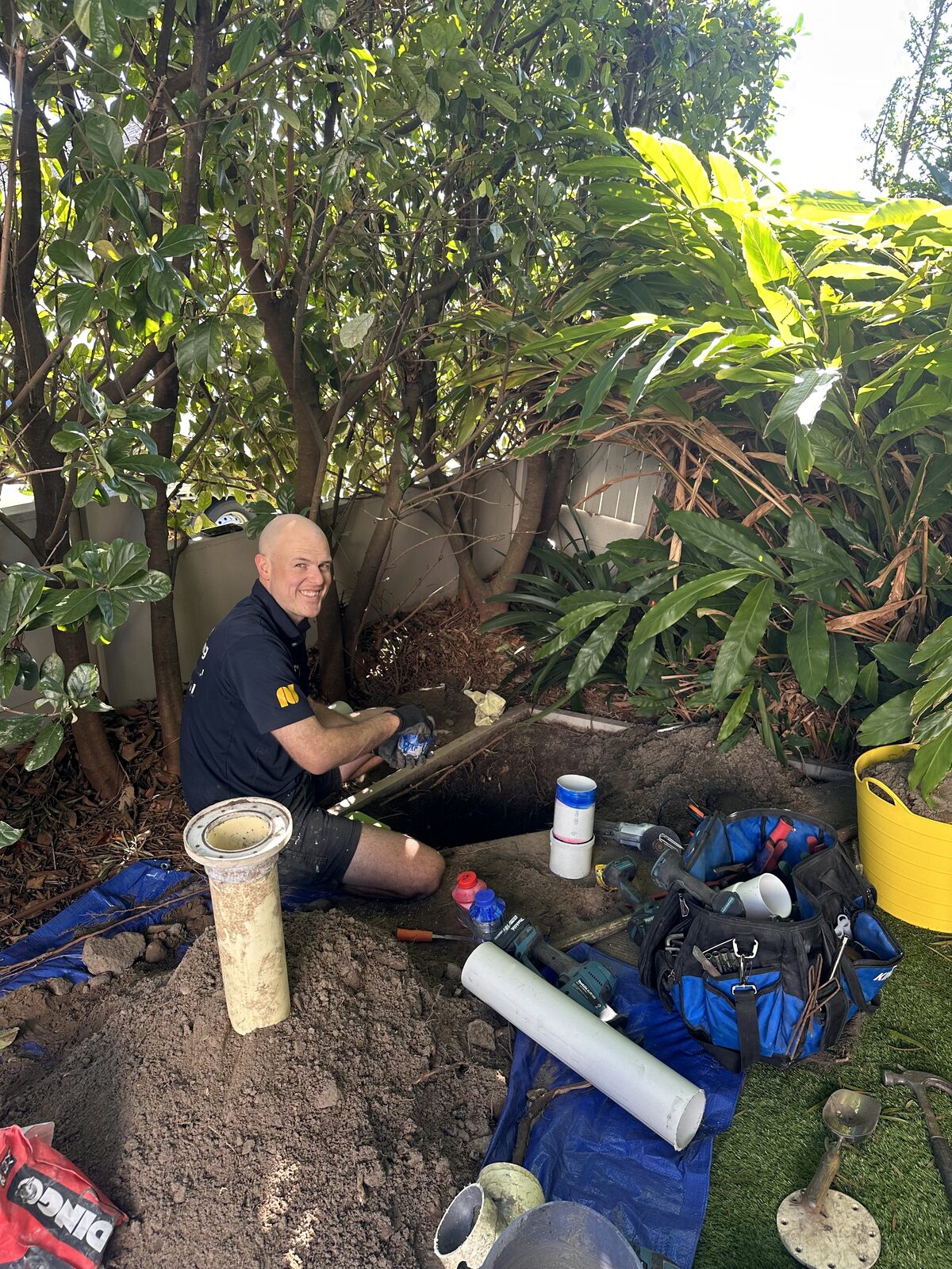 Greg from Norton Plumbing fitting new PVC pipe into an excavated drain trench in a South Coogee garden bed, surrounded by tools and old terracotta pipe sections
