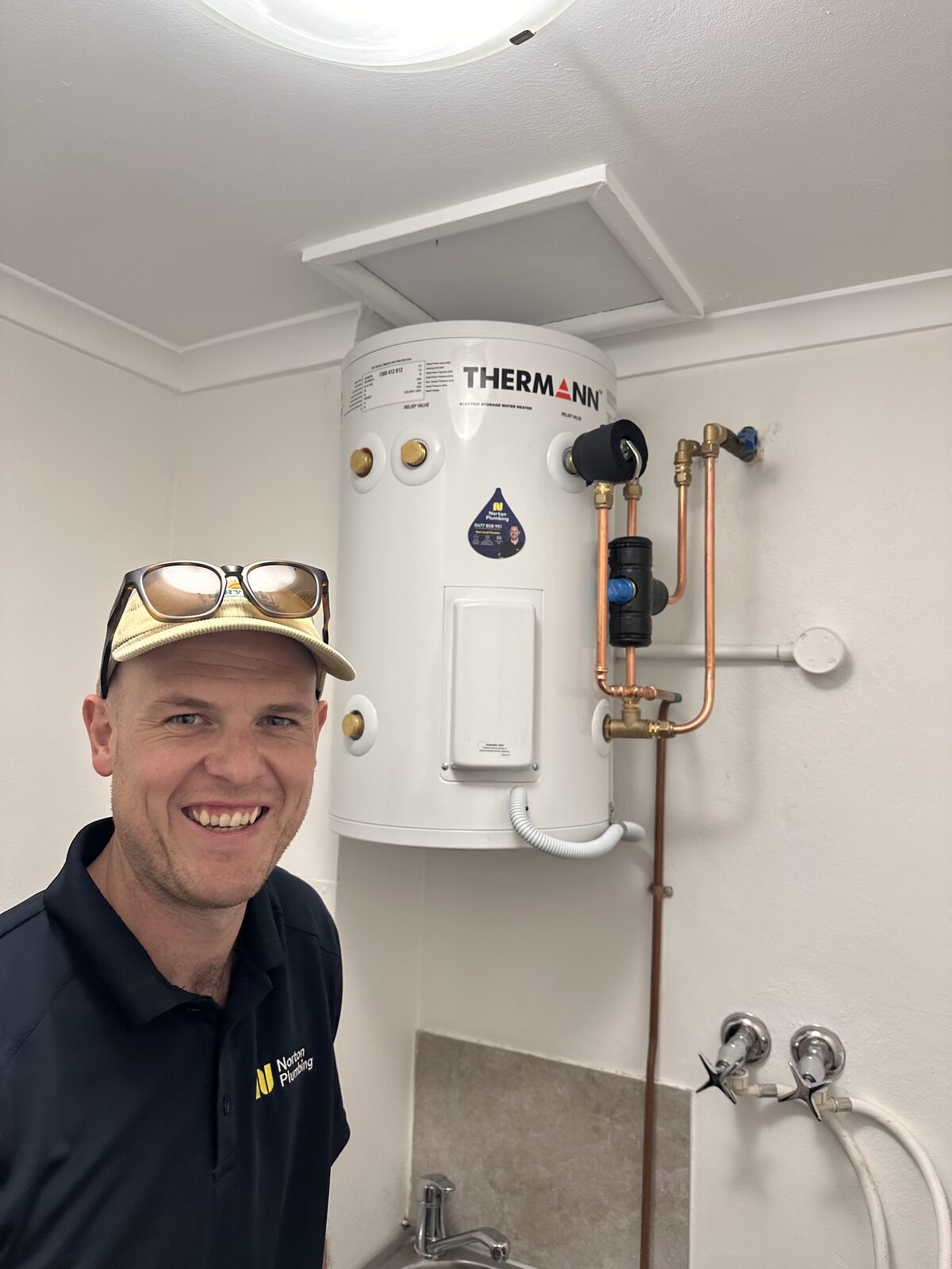 Greg from Norton Plumbing beside a newly installed Thermann electric storage hot water system mounted high on a laundry wall in Coogee