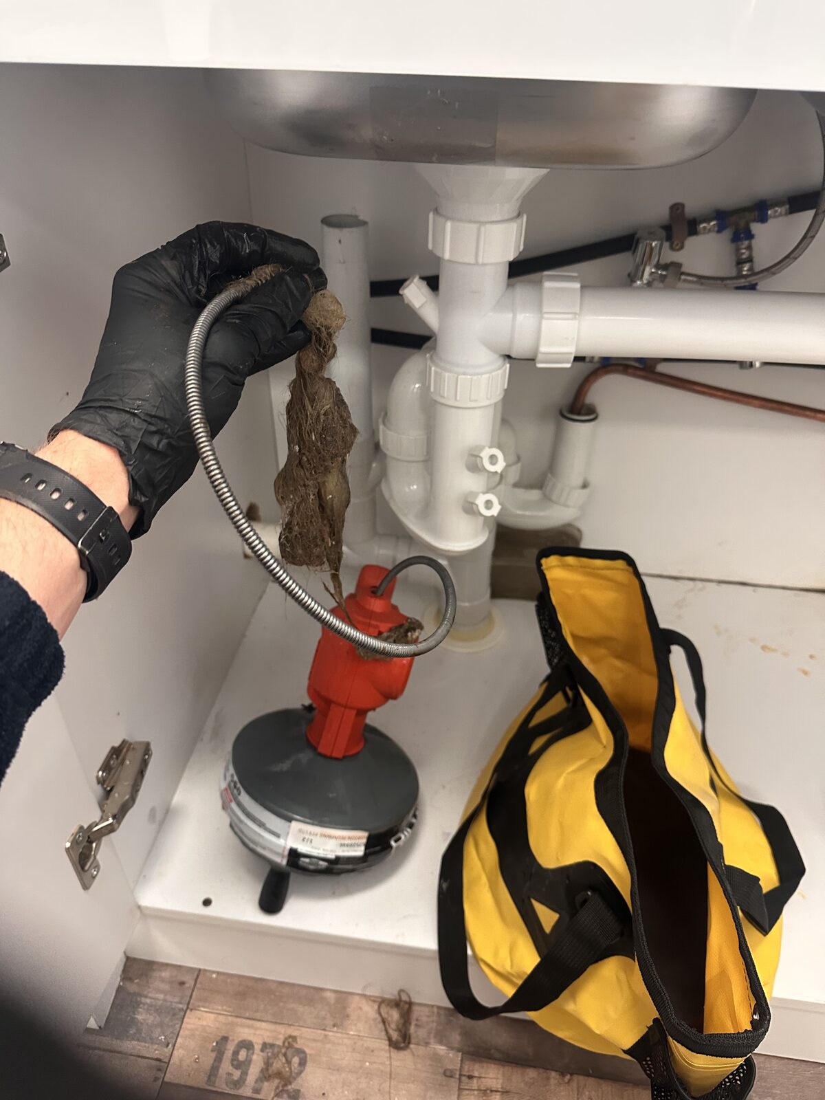 A gloved hand holding a large rope of matted hair pulled from a blocked drain at a Maroubra hairdressing salon, with a drain machine on the floor below