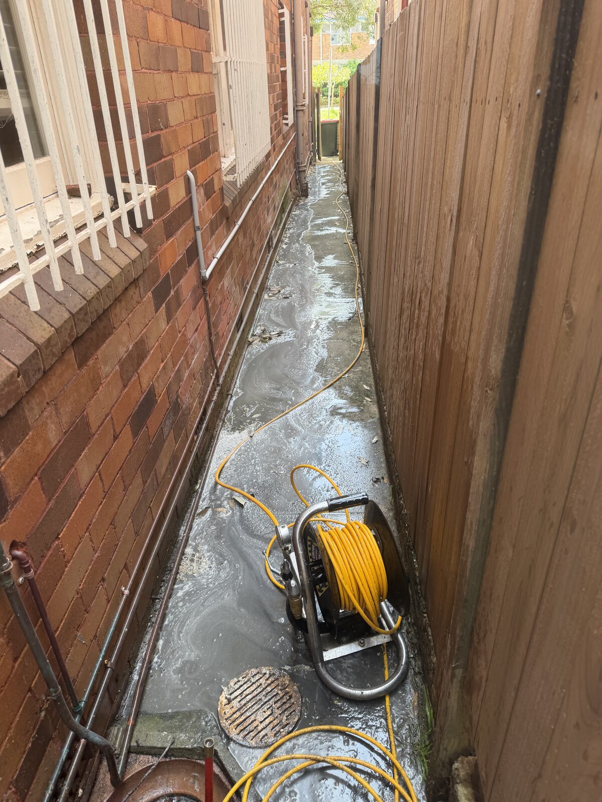 Norton Plumbing drain jetter set up beside an overflowing sewer gully in the narrow side passage of a red brick strata block in Bondi, with wastewater spread across the concrete