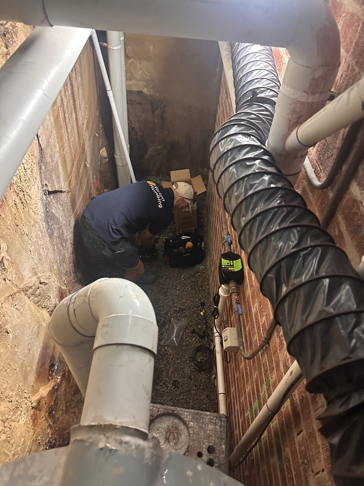 Looking down into the gravel-floored subfloor void of a Dover Heights house where a Norton Plumbing plumber is working on the rainwater pump, with stormwater pipes and air conditioning ducting overhead