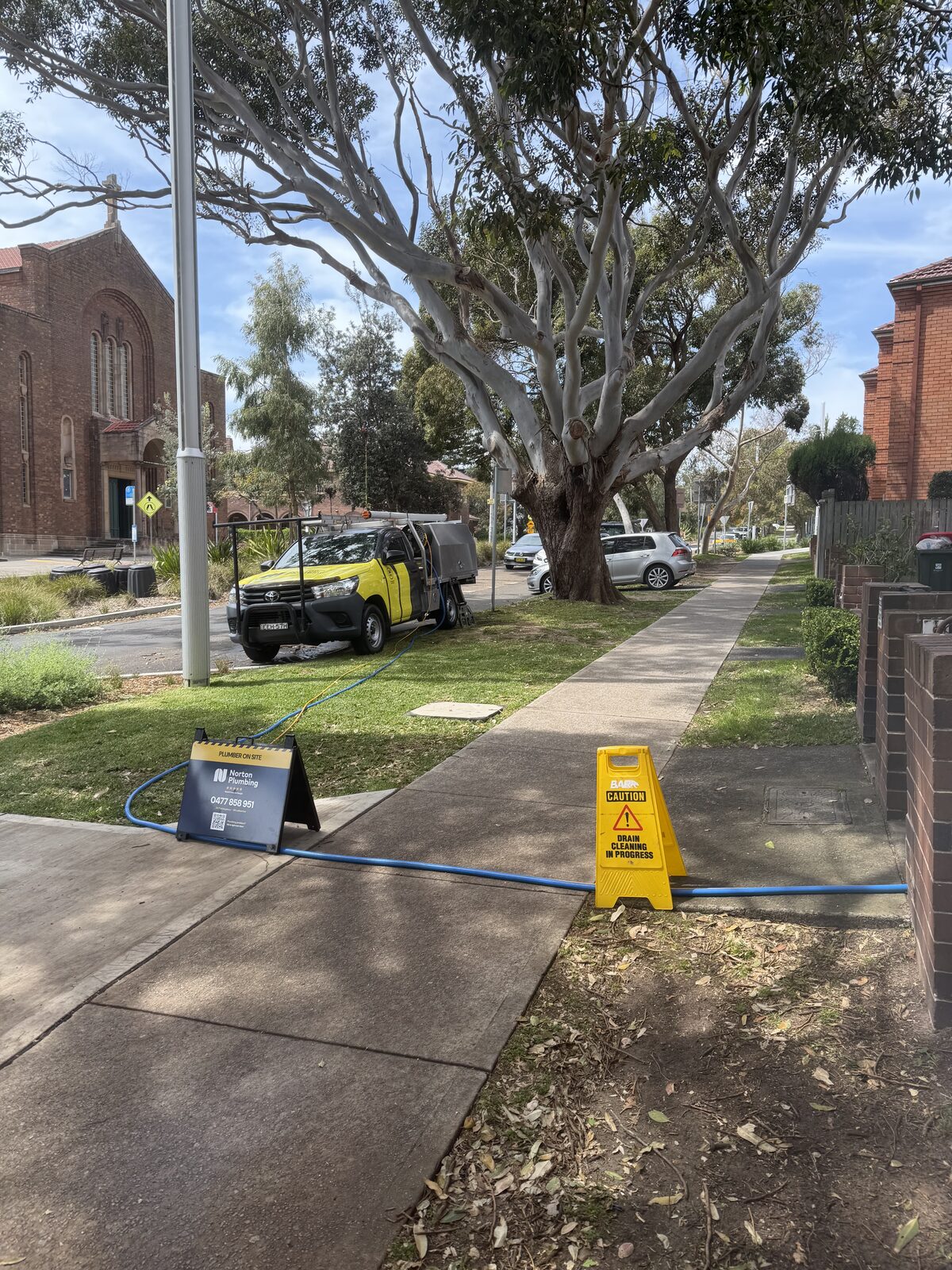 Norton Plumbing ute parked on a Bondi street with a plumber on site sign and yellow safety sign on the footpath, and a hose running across the nature strip to a strata block