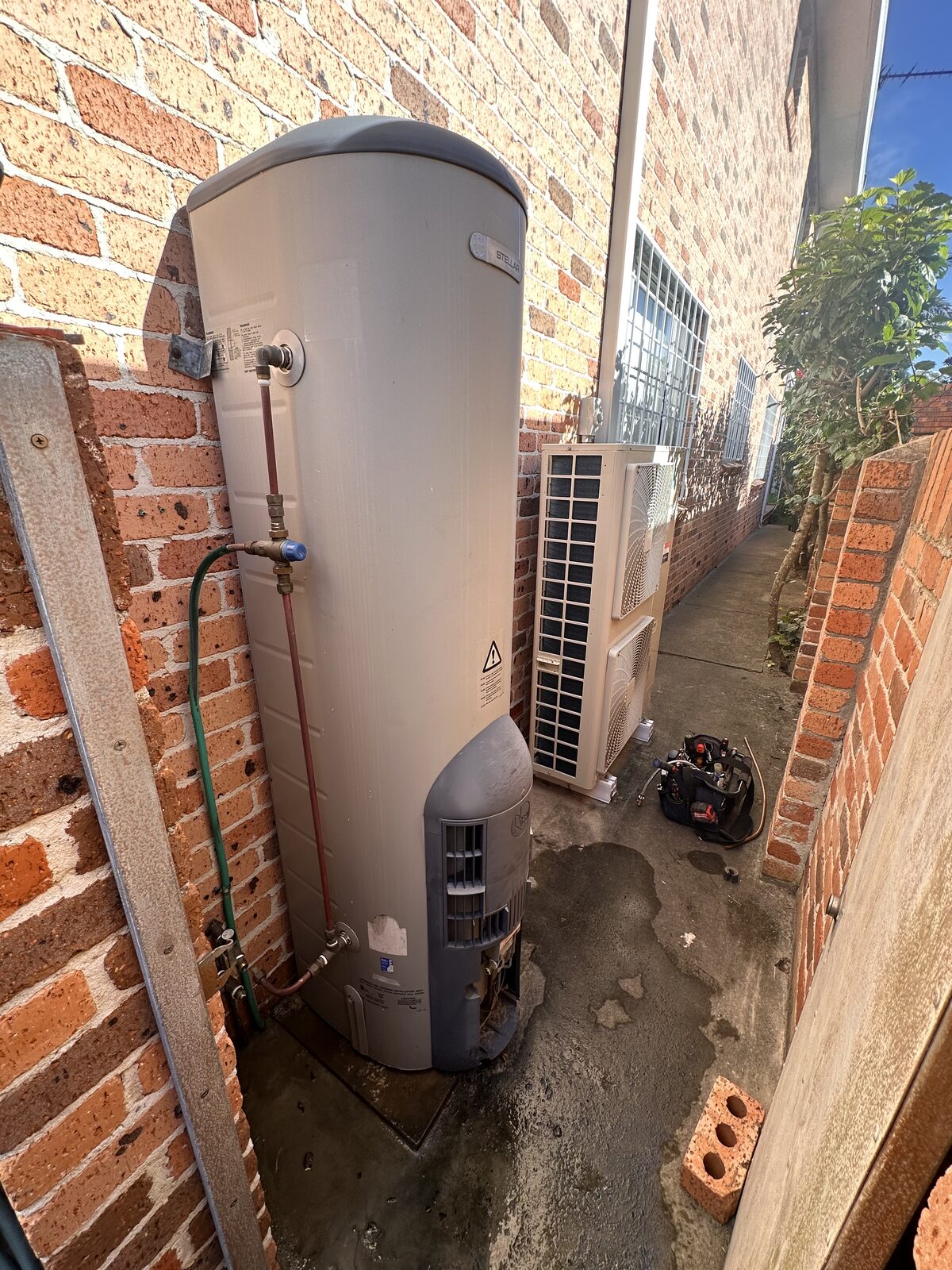 Old leaking gas hot water system standing against a brick wall in a Maroubra side passage, with water pooling at the base