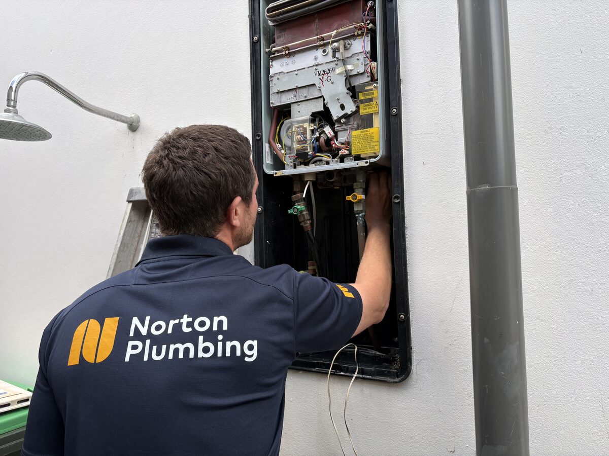 Adam from Norton Plumbing working on the old Rinnai Infinity 26 gas instantaneous hot water system with its cover off in a wall recess box at a Clovelly house
