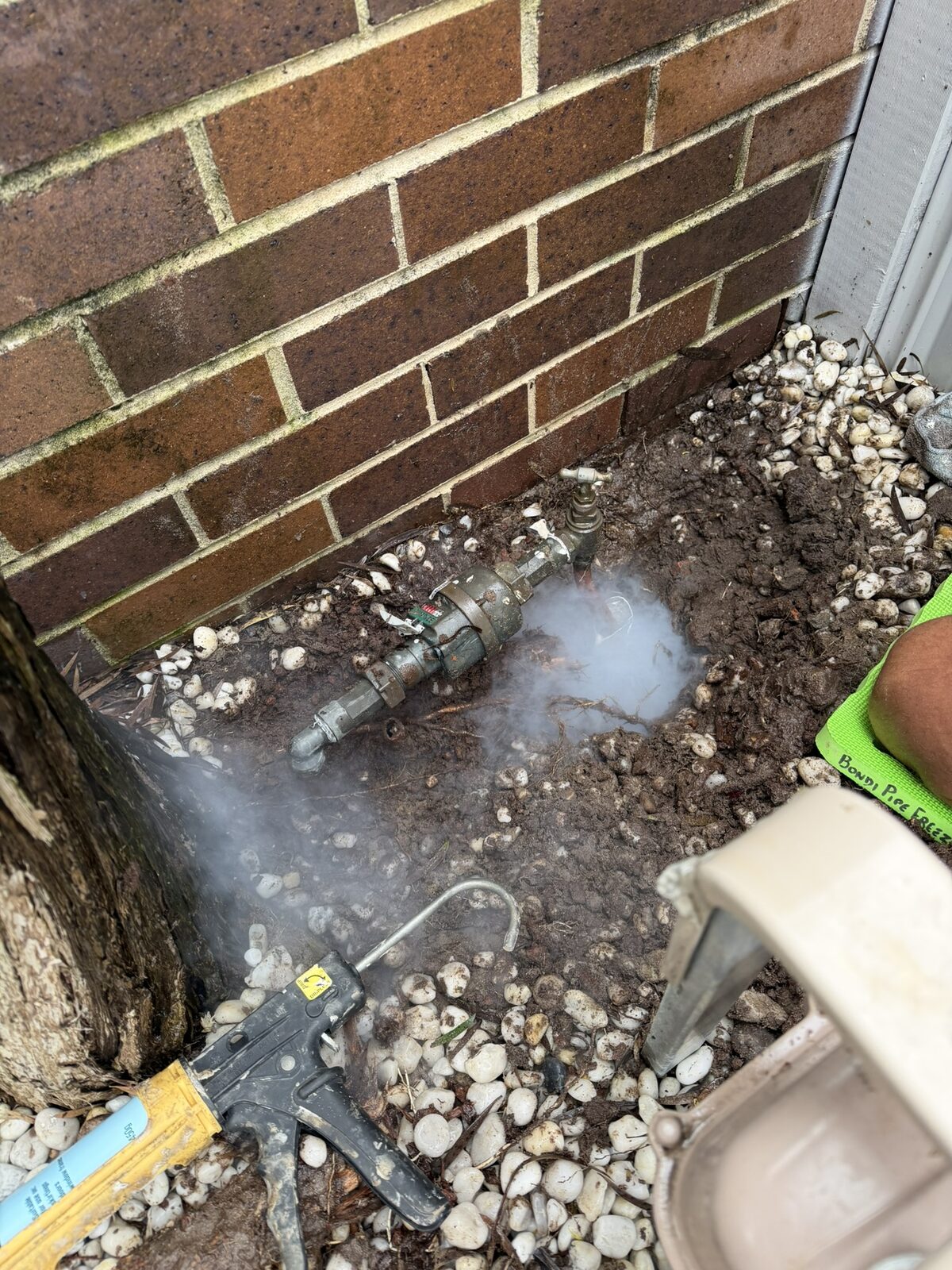 Pipe freezing kit fogging over the copper riser at a leaking water meter tap in a Maroubra garden bed, with the brass meter and brick wall behind