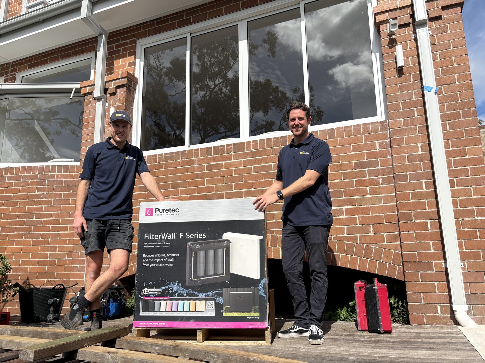 A boxed Puretec FilterWall F Series whole-house water filter on a pallet outside a red brick Randwick home, with Adam Norton and Greg from Norton Plumbing either side of it