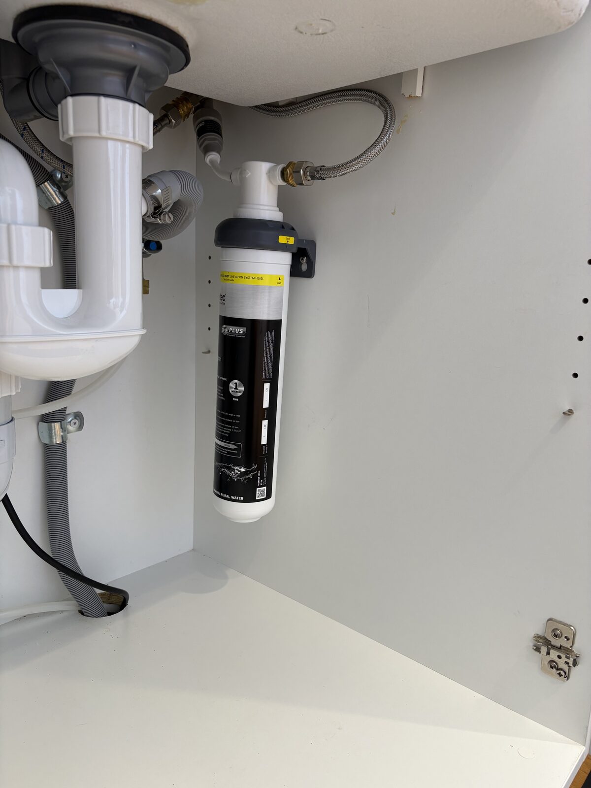 Close-up of a Puretec PureMix Z7 inline water filter mounted on the cabinet wall under a Coogee kitchen sink, with the braided cold water flexi hose connected to the filter head