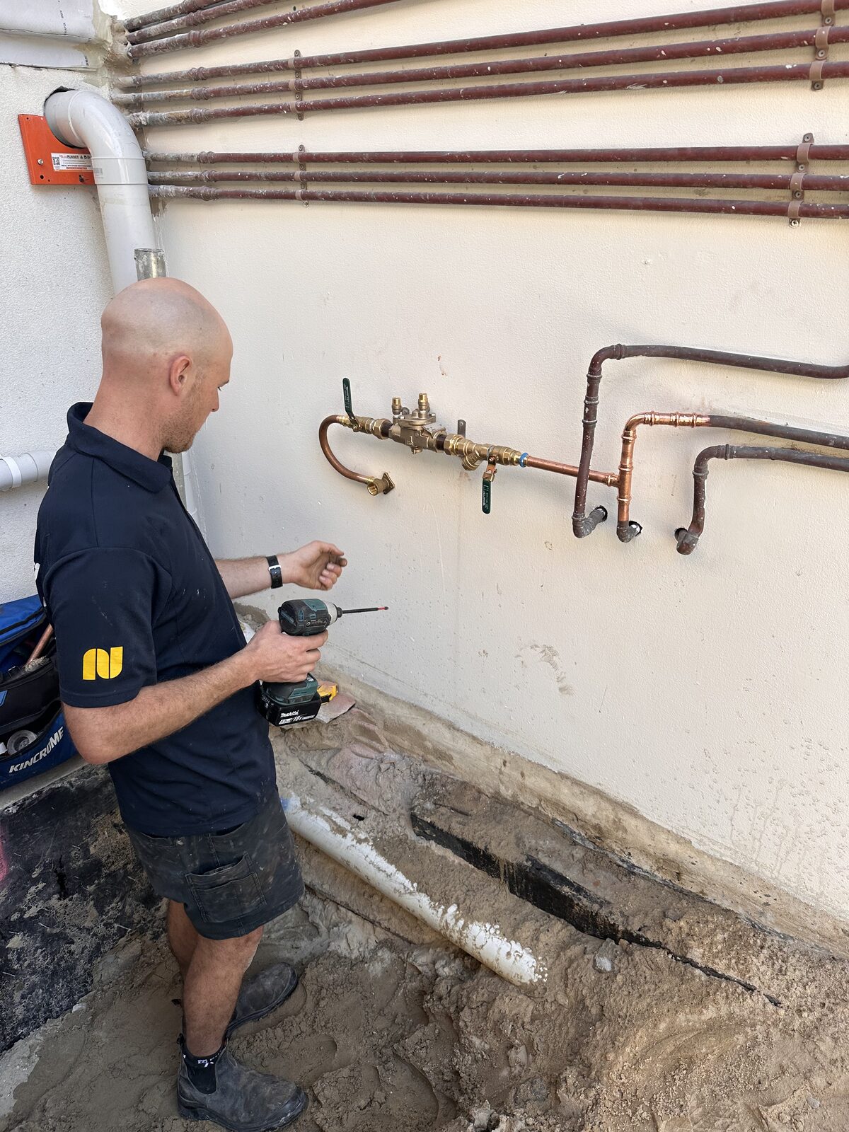 Wide view of a Bondi building's external wall showing the new RPZ backflow prevention device mounted below the original water service pipework, with a trench open along the base of the wall