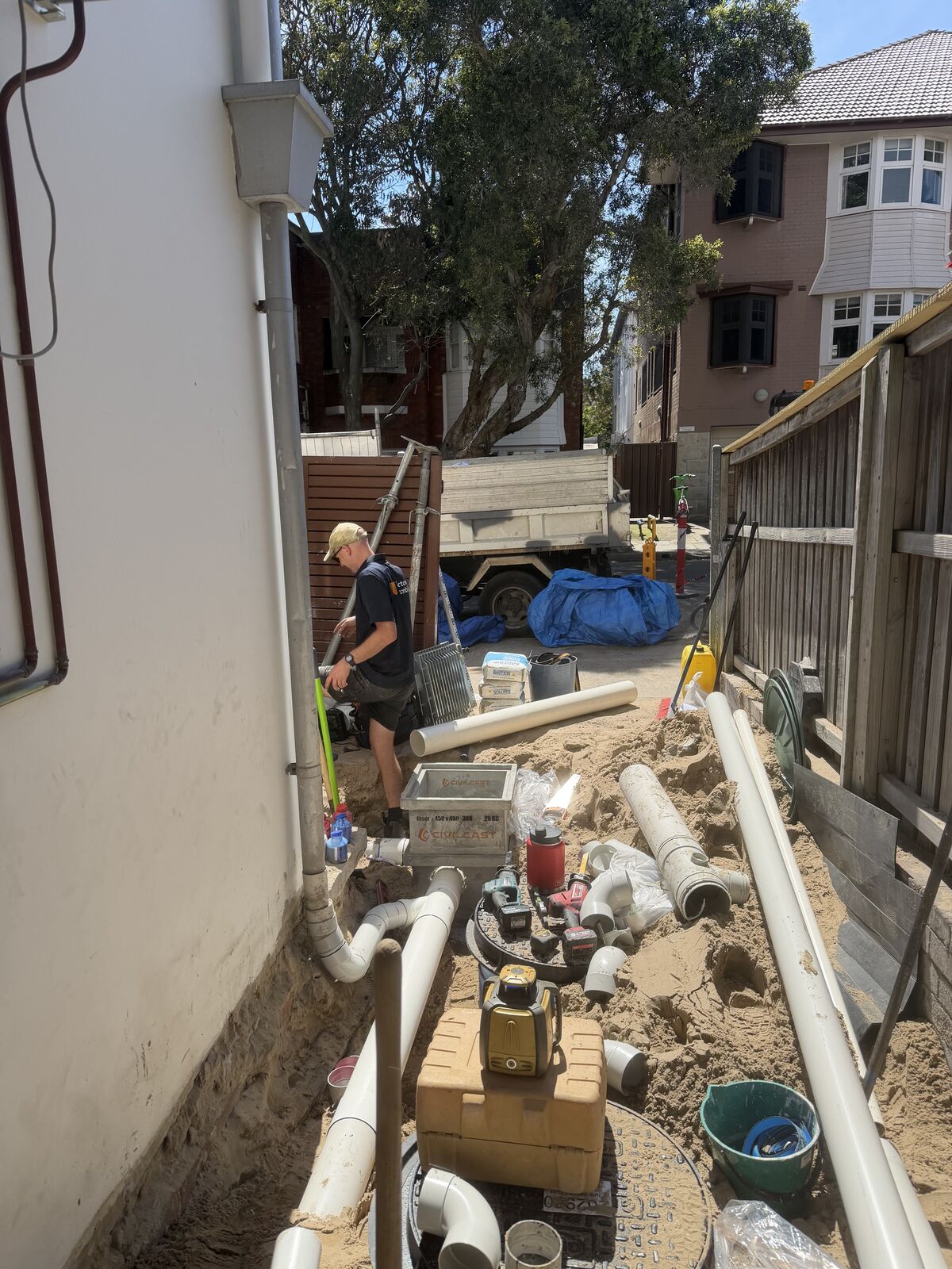 A narrow side passage at a Bondi home dug out along its full length, with new PVC stormwater pipe laid in the sandy trench beside the house and Greg working at the far end