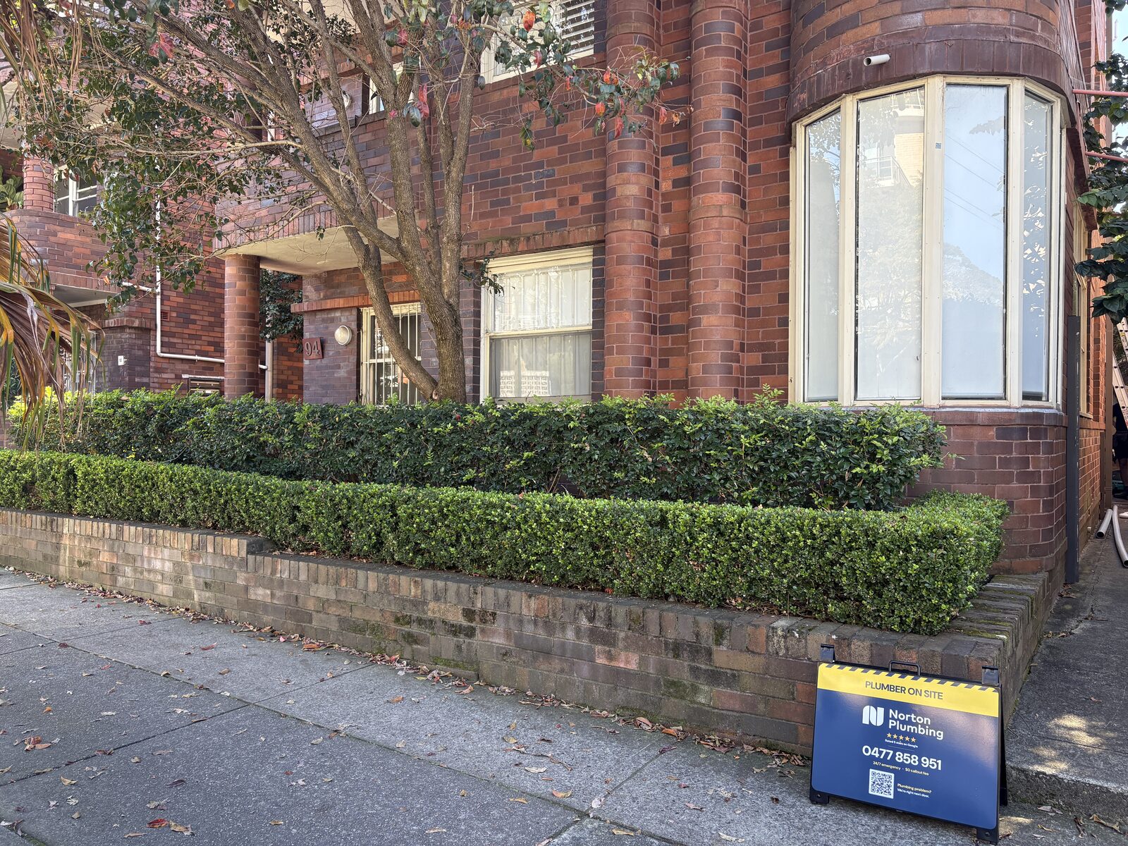A three storey interwar red brick strata apartment block on Coogee Bay Road, Coogee, with curved bay windows and a Norton Plumbing plumber on site sign on the footpath
