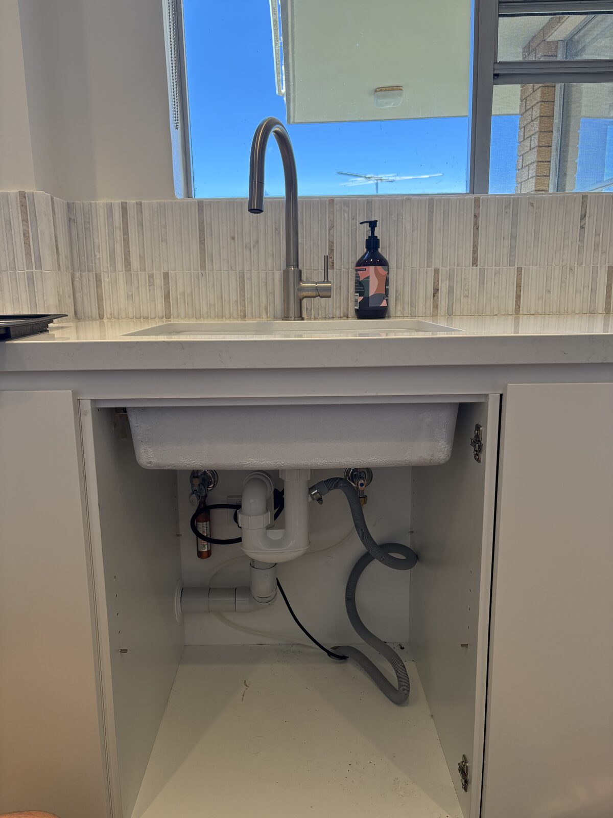 The sink cupboard of a renovated Coogee apartment kitchen before an under-sink water filter was installed, showing the existing trap, waste and flexible supply hoses
